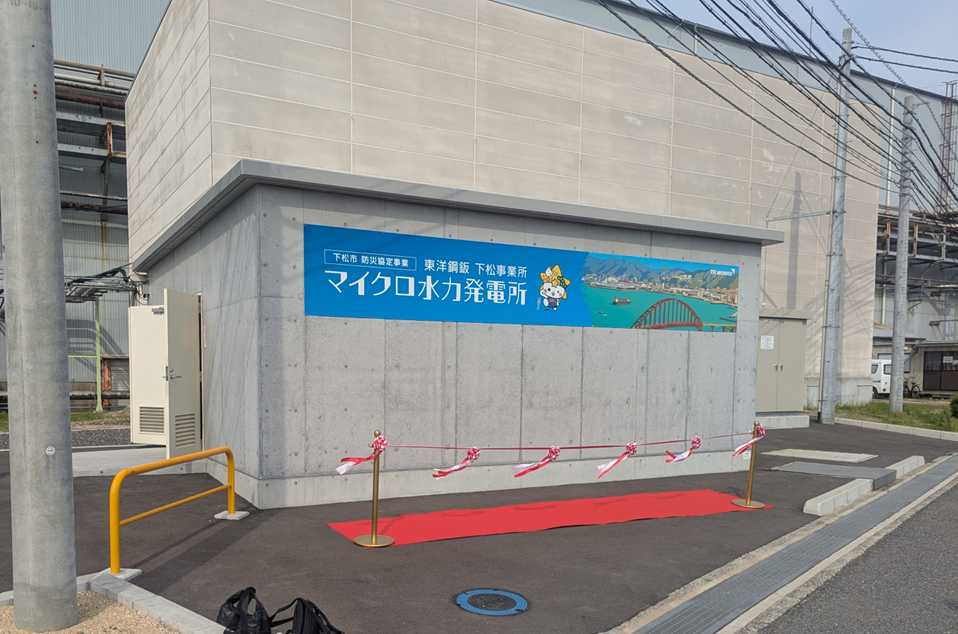 東洋鋼鈑株式会社下松事業所内に建設された「下松事業所マイクロ水力発電所」の外観。コンクリート造りの建屋壁面には、青色を基調とした鮮やかな看板が設置されている。看板には「下松市 防災協定事業」「東洋鋼鈑 下松事業所」「マイクロ水力発電所」の文字に加え、下松市の公式マスコットキャラクター「くだまる」や地域の風景が描かれている。