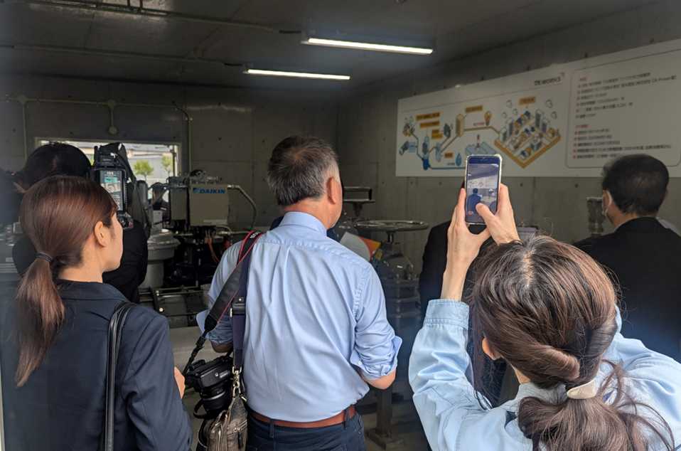 東洋鋼鈑下松事業所マイクロ水力発電所開所式における、メディア取材の様子。コンクリート造りの発電所建屋内にて、報道関係者や関係者が多数詰めかけている。中央にはプロ仕様の一眼レフカメラを肩にかけた報道関係者、右側にはスマートフォンで建屋内や奥に鎮座する「DAIKIN」ロゴ入りの縦型発電機を撮影する出席者の姿があり、本プロジェクトへの注目の高さがうかがえる。