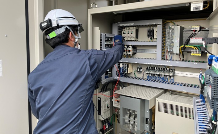 技術者が、屋外に設置されたマイクロ水力発電システム(小水力発電システム)の制御盤を点検している様子。精密な配線とメンテナンスを徹底