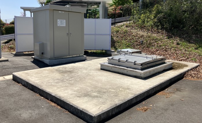 藤岡市南山配水池マイクロ水力発電所の屋外設置設備。グレーのシステム制御盤と、地下ピットへアクセスするための金属製ハッチが、限られた敷地にコンパクトに配置されている様子。既存施設の構造を変えずに導入可能であることを示している。