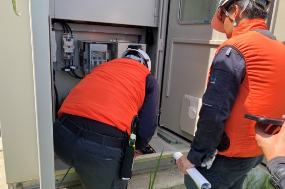オレンジ色の作業服を着たメンテナンススタッフが屋外システム制御盤を開けて設備点検を行っている様子