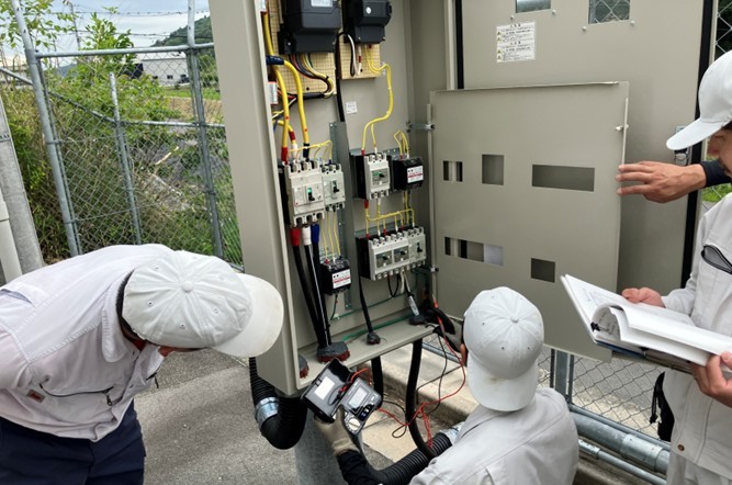 屋外の配電盤を開け、測定器を用いて電気系統の定期点検と数値確認を行う作業員たち。DK-Powerは、発電設備の点検を定期的に実施し、安定稼働を維持しています。
